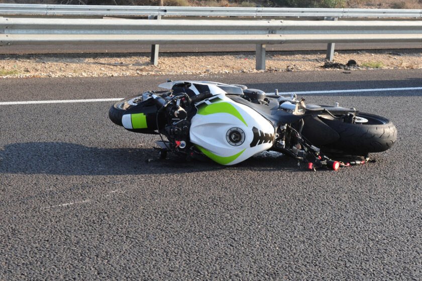 A white and green motorcycle lies on its side on an asphalt road next to a highway guardrail. The bike shows signs of damage, suggesting a recent accident. The background includes dry grass and a metal barrier.