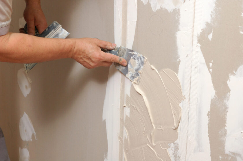 A person is applying joint compound to drywall using a putty knife, smoothing it over the seams and screw heads. The wall appears partially finished, with some areas already coated and others still bare.
