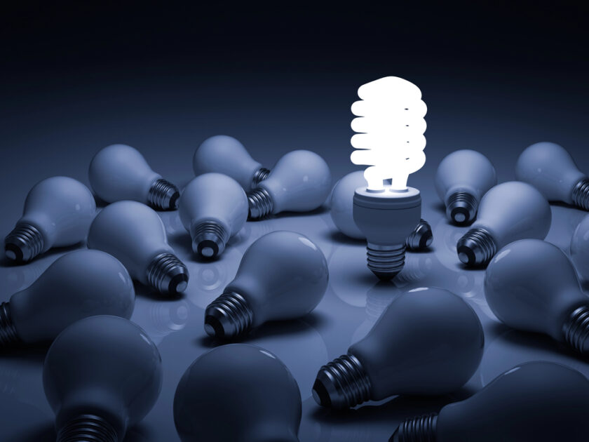A lit spiral fluorescent bulb stands upright among numerous unlit incandescent light bulbs lying on a dark surface. The glowing bulb creates a striking contrast against the surrounding dimness.