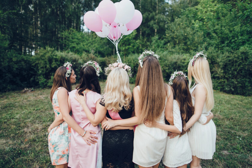 Six women in dresses and floral crowns stand with their backs to the camera, embracing each other. They are outdoors on grass, surrounded by greenery. One holds a bunch of pink and white balloons.