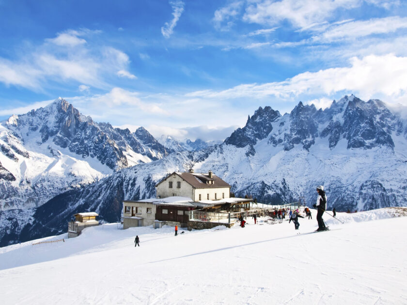 A snow-covered mountain slope with several skiers and snowboarders. In the background, theres a lodge surrounded by majestic, jagged mountains under a clear blue sky. Some clouds hover over the peaks.