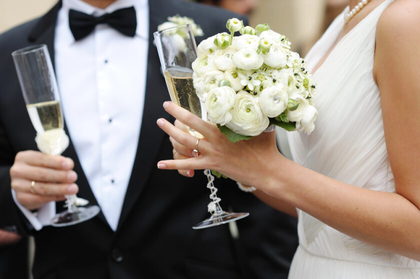 Bride in white dress holding a bouquet of white flowers and a champagne flute, standing next to a groom in a black tuxedo with a bow tie, also holding a champagne flute.