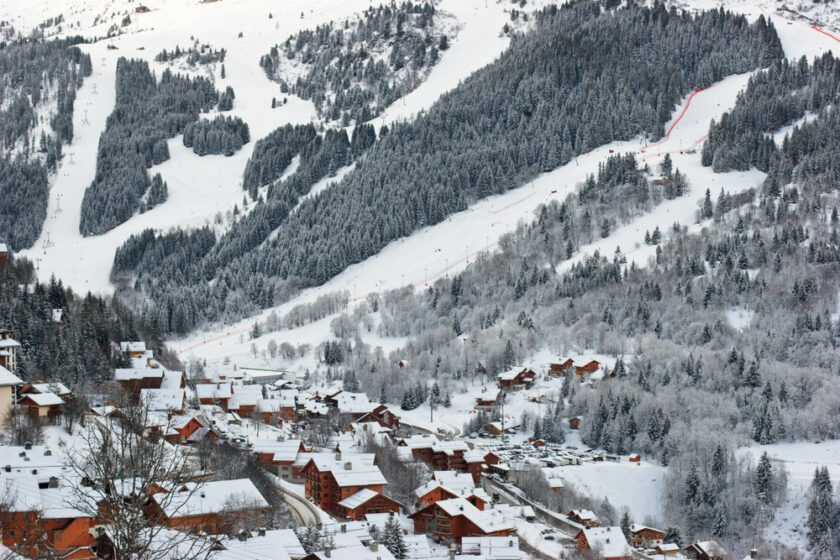 Snowy alpine village with wooden chalets scattered across the landscape. Pine trees and snow-covered slopes dominate the background, indicating a ski area. The scene is peaceful and wintry, capturing the charm of a mountain retreat.