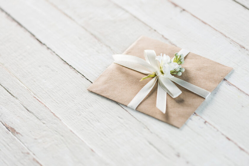 A brown envelope adorned with a white ribbon and small white flowers lies on a light wooden surface. The arrangement gives a delicate and elegant appearance.