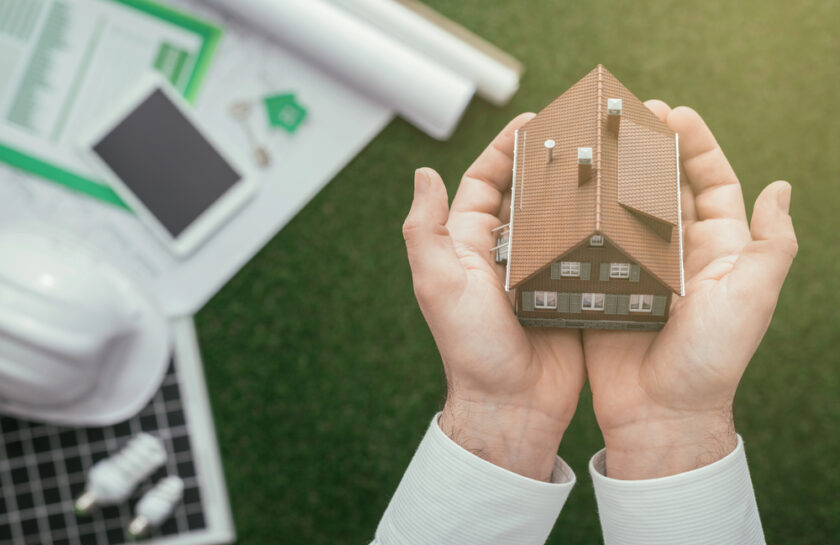 Hands holding a small model house. Nearby are architectural plans, a smartphone, a white hard hat, and two light bulbs on a green surface, suggesting a theme of sustainability and planning in real estate or construction.