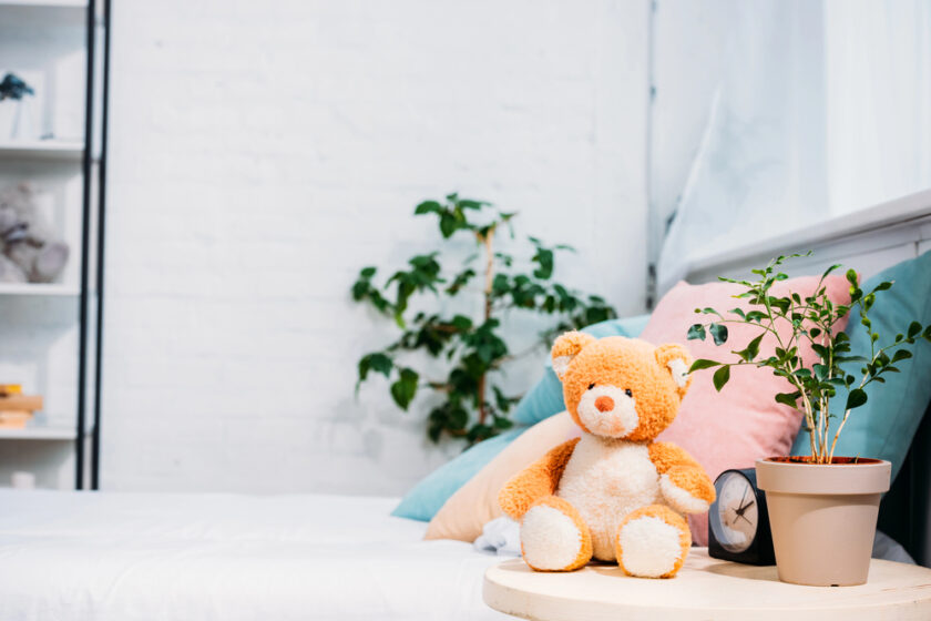 A cozy bedroom with a neatly made bed featuring pastel cushions. A teddy bear sits on a wooden nightstand beside a potted plant and a clock. Soft lighting illuminates the white brick wall and a small plant in the corner.