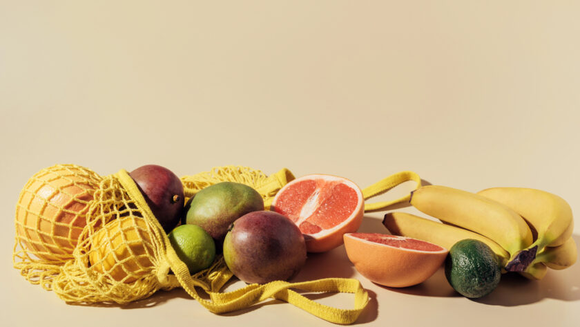 A yellow net bag with mangoes, limes, and passion fruits spills over a beige surface. Next to it are halved grapefruits, a bunch of bananas, and an avocado. The lighting is bright and warm, highlighting the fruits colors and textures.