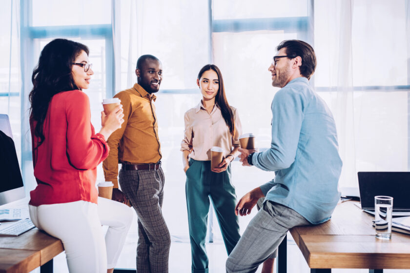 Four colleagues are having a casual meeting in an office. Three are standing, holding coffee cups, and one is sitting on a desk. They are engaged in conversation, and natural light is streaming through large windows behind them.