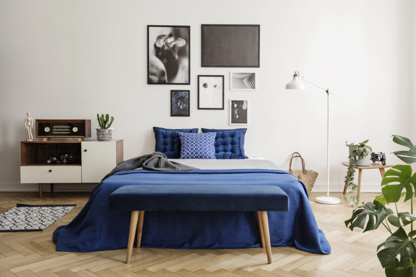 A modern bedroom with a bed covered in blue linens and multiple pillows. A gallery wall features black and white photos. Theres a wooden bench at the foot of the bed, a floor lamp, a sideboard, plants, and a decorative rug on a wooden floor.