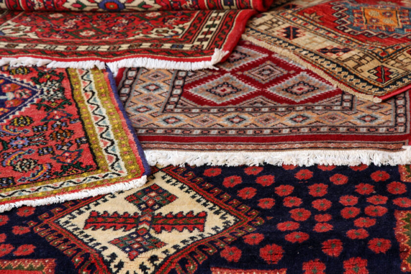 A display of colorful, intricately patterned Persian rugs overlapping each other. The patterns include geometric shapes and floral designs in red, blue, cream, and green hues, with fringed edges visible.