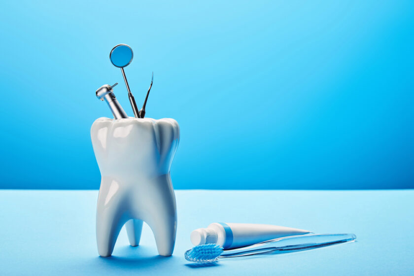 A large model of a tooth holds dental tools, including a mirror and explorer. Nearby, a toothbrush and toothpaste lie on a bright blue surface, with a blue background.