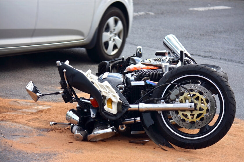A motorcycle lies on its side on the road with visible damage, next to a pool of sand. Part of a white car is visible nearby, suggesting an accident scene. The background is slightly blurred, focusing on the motorcycle and the cars wheel.