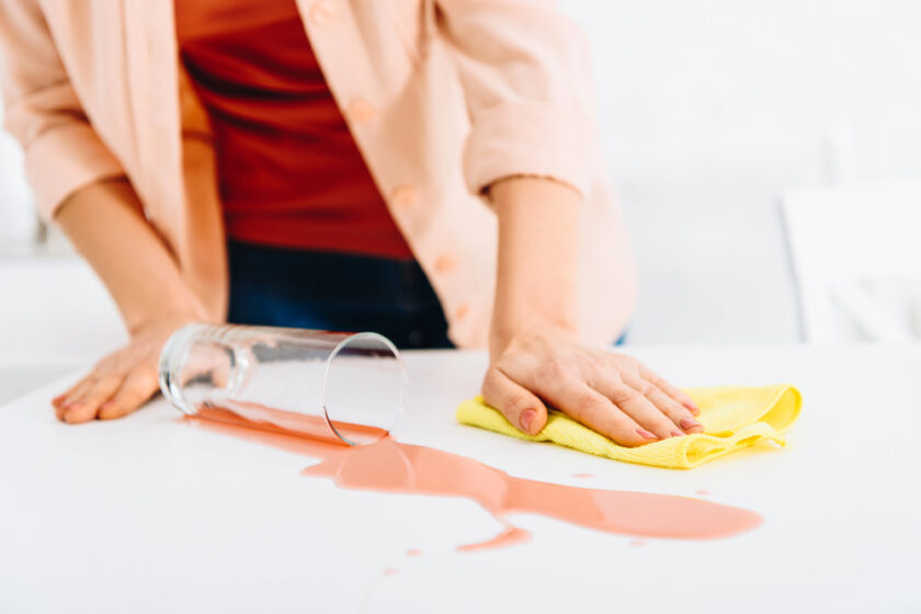 A person is wiping up spilled juice from a white table with a yellow cloth. The juice is pink and the glass is tipped over. The person is wearing an orange shirt and red top.