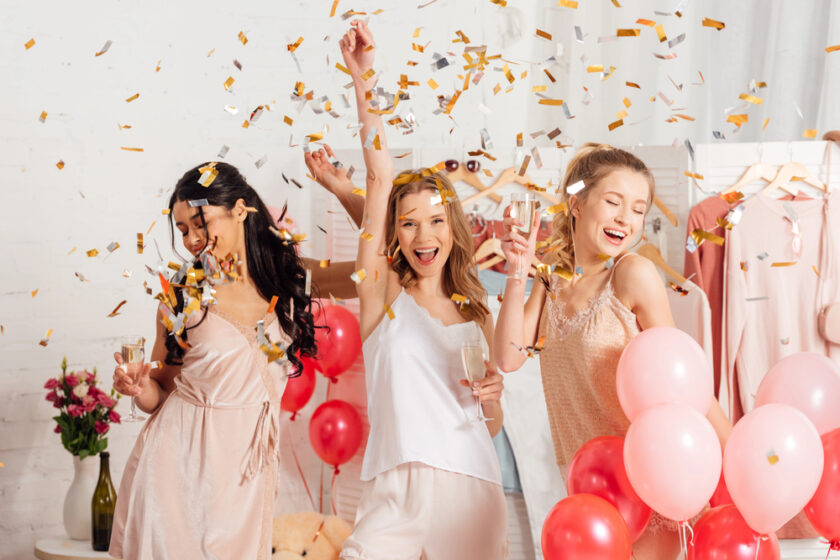 Three women in pajamas joyfully celebrate amidst falling confetti and balloons. They hold champagne glasses and smile brightly. The background features red and pink balloons and a vase with flowers, creating a festive atmosphere.