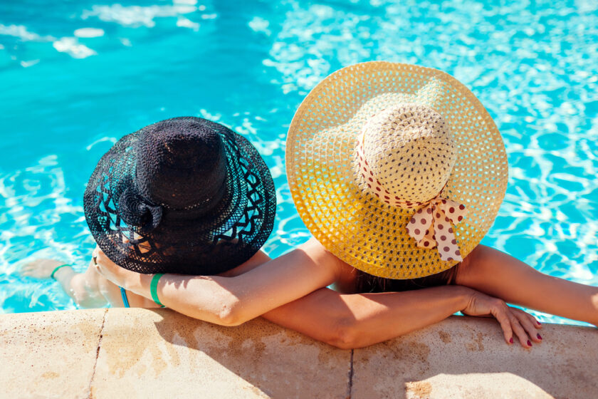 Two people wearing wide-brimmed hats, one black and one yellow with polka dots, are relaxing at the edge of a swimming pool. They have their backs to the camera, arms resting on the poolside, enjoying the sunny day.