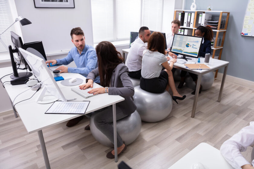 People are working in an office. Some are seated on exercise balls around desks with computers and charts. The office has bright windows, light-colored walls, and wooden flooring.