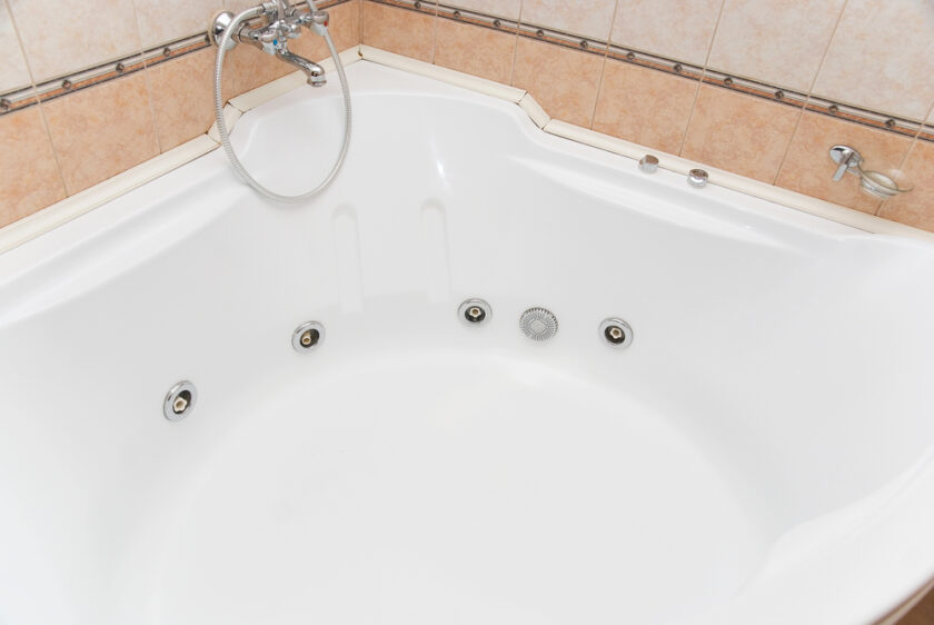 A white, empty corner bathtub with six round jets is shown. It features a silver faucet and a removable handheld shower head. The tub is set against beige tiled walls with decorative trim.