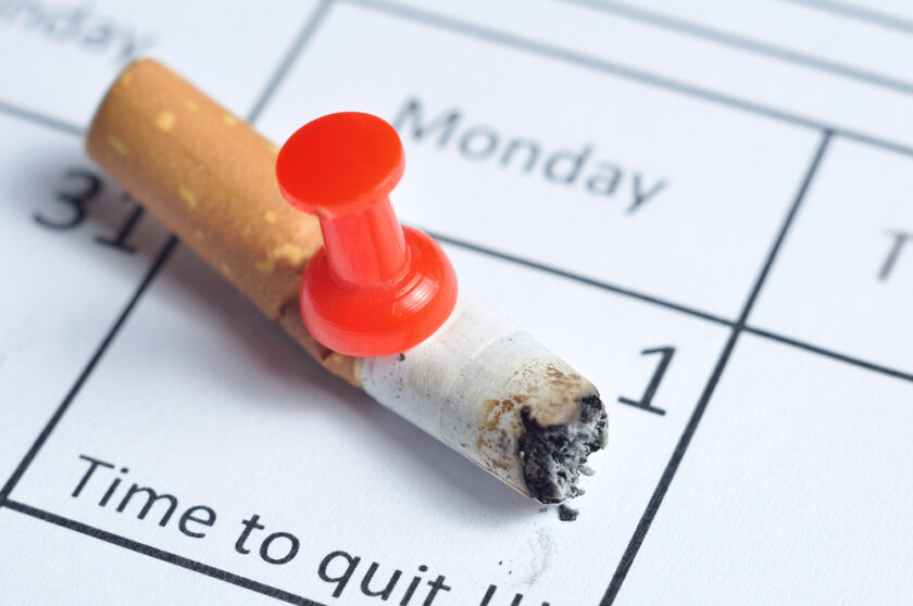 A cigarette butt with a red pushpin through it rests on a calendar, highlighting a day labeled Time to quit. The image symbolizes quitting smoking and setting a specific date to stop.