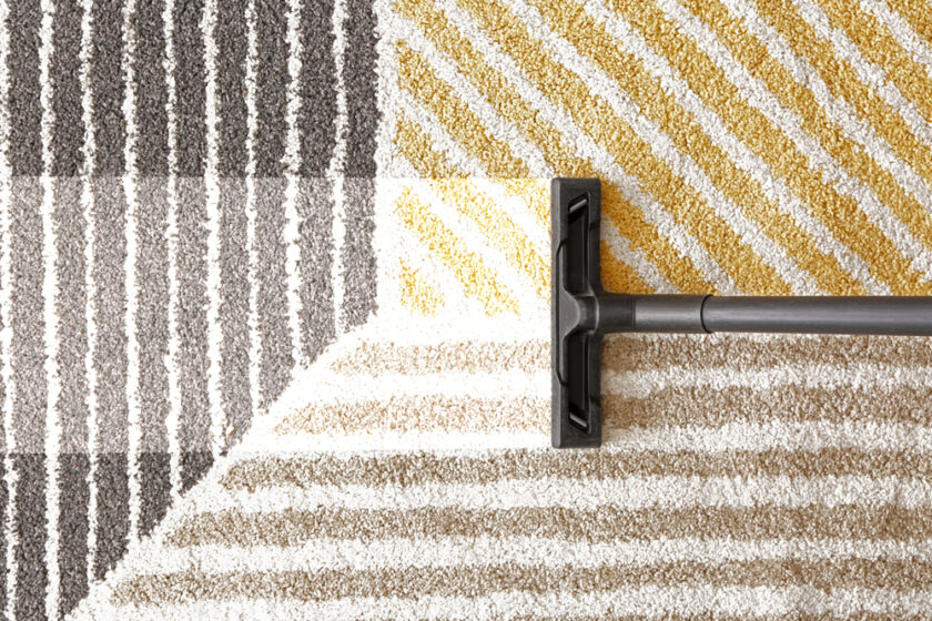 A vacuum cleaner head on a textured carpet with diagonal stripes in gray, yellow, and tan shades. The carpet features a geometric pattern with sections in various colors, creating a contrast between clean and unclean areas.