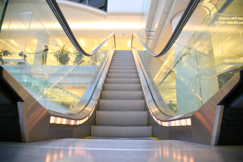 Modern indoor escalator with glass sides, leading upwards. Bright, reflective surfaces and overhead lights illuminate the area. Surrounding architecture features clean lines and a few potted plants.