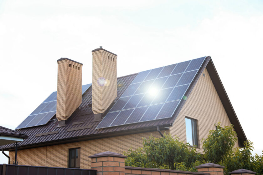A brick house with two chimneys features solar panels installed on its sloped roof. The sky is clear, and the sun is reflecting off the panels. Green bushes are visible in the foreground.