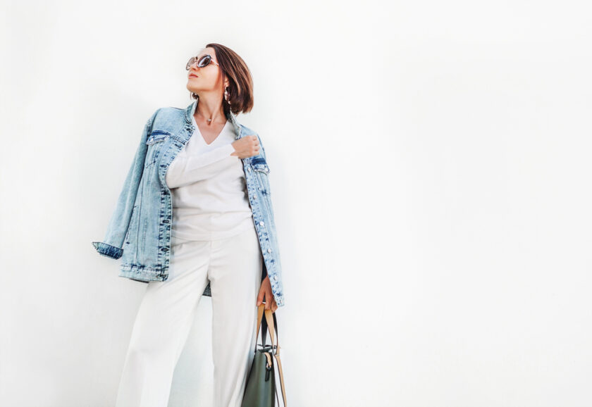 A person in sunglasses poses confidently against a white background, wearing a denim jacket over a white top and pants. They hold a handbag in one hand and gaze upward.