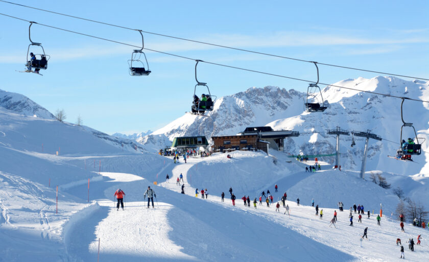 A snowy ski resort with numerous skiers on the slopes and several people riding ski lifts. In the background, a chalet sits amid majestic snow-covered mountains under a clear blue sky.