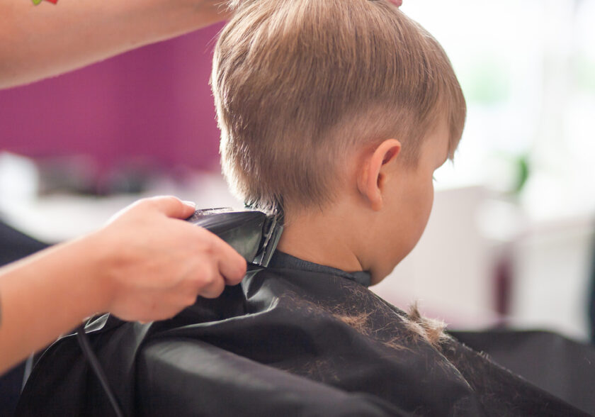 A child is getting a haircut. The hairstylist is using clippers to trim the back of the childs head. The child is wearing a black cape, and some hair clippings can be seen on it. The background is blurred.