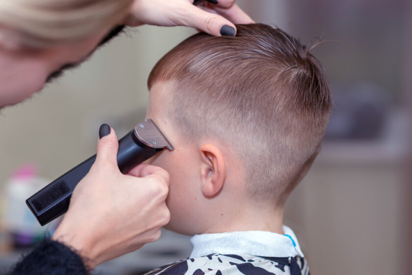 A stylist uses clippers to trim the hair of a young boy, creating a neat, short haircut. The boy sits calmly in a barber chair, while the stylist focuses on the task. Nearby, haircutting tools and a spray bottle are visible.