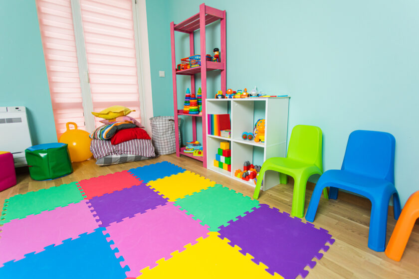 A colorful playroom with a vibrant foam mat on the floor. Shelves hold toys and blocks, and two bright chairs, one green and one blue, are nearby. A stack of pillows sits by a window with blinds, adding to the playful atmosphere.