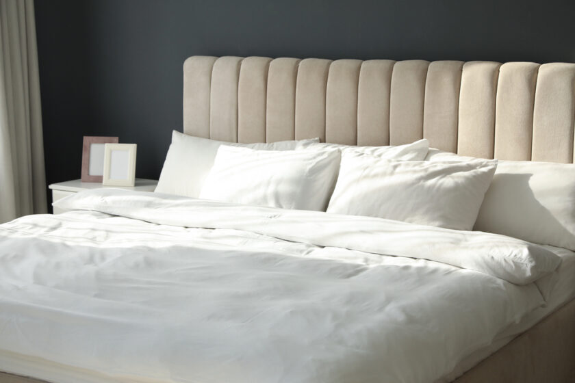 A neatly made bed with a beige upholstered headboard and white bedding. Two white pillows are placed against the headboard. On the bedside table are two empty picture frames. Sunlight streams through a window off-screen.