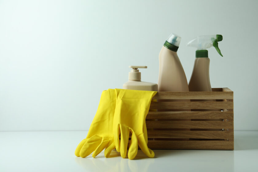 A wooden crate holds various beige cleaning bottles and a spray bottle. Yellow rubber gloves are draped over the side of the crate. The setting is minimalistic with a plain white background.