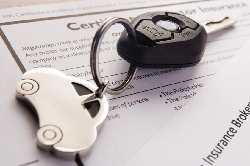 Car keys with a car-shaped keychain rest on top of car insurance documents. The keys have a black remote with buttons, and the paperwork is partially visible, suggesting topics related to vehicle insurance.