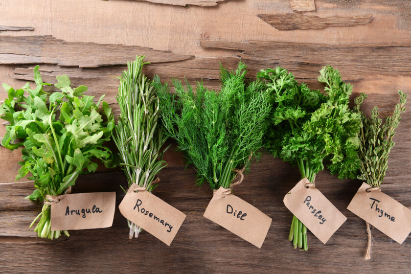Five bundles of fresh herbs on a wooden surface, each with a brown paper tag labeled: arugula, rosemary, dill, parsley, and thyme.