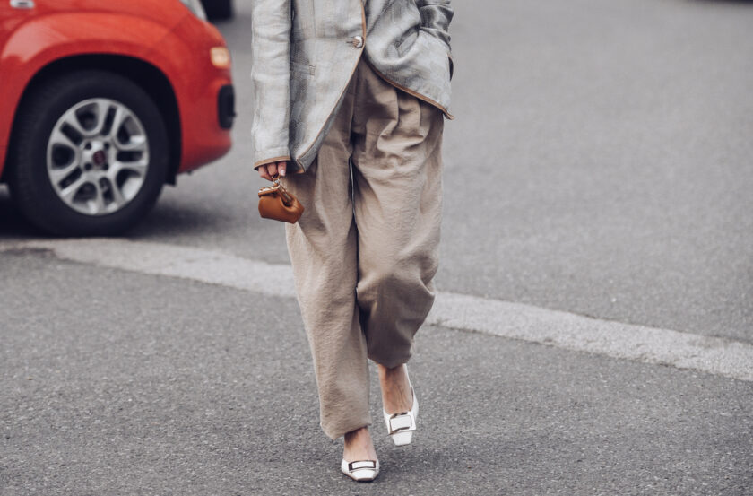 Person walking on a street, wearing a gray plaid blazer, loose beige trousers, and white shoes. They are holding a small brown handbag. A red car is parked in the background.