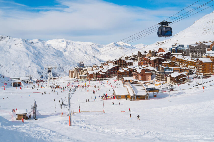 Scenic view of a snowy ski resort with skiers on the slopes, ski lifts, and a cable car overhead. Chalets and buildings are nestled in the background, surrounded by snow-covered mountains under a clear blue sky.