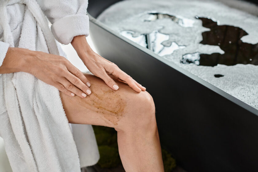 A person in a white bathrobe applying a scrub to their knee in front of a filled bathtub. The focus is on their hands and the exfoliating product being used on the skin.