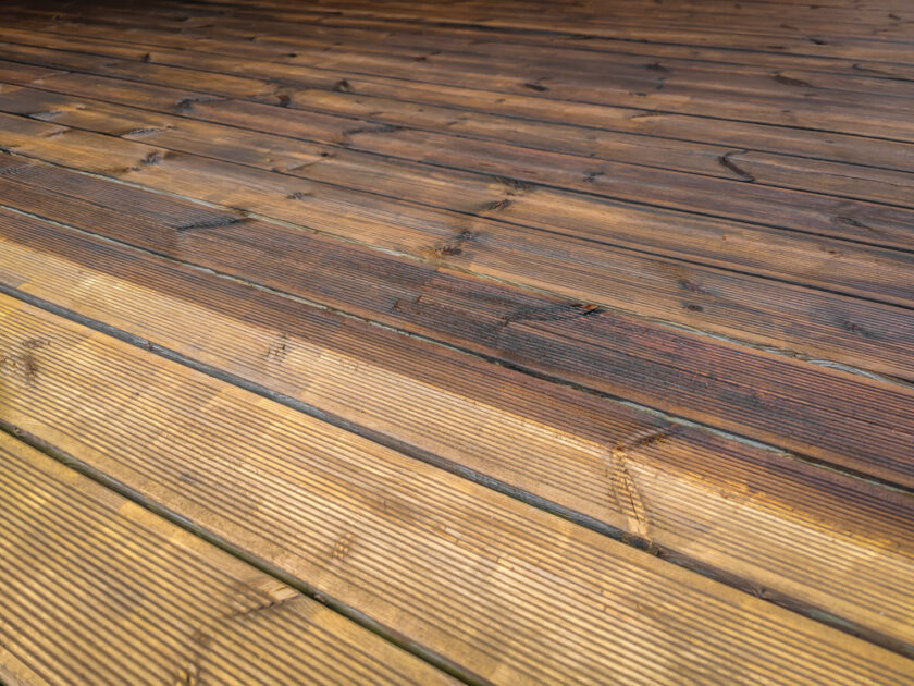 Close-up of a wooden deck with horizontal planks. The wood shows variations in color, from light to dark brown, highlighting the natural grain and texture. Subtle sunlight casts shadows, enhancing the texture and pattern.