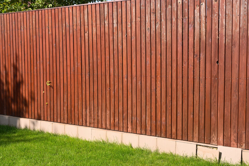 A tall wooden fence with vertical planks painted red-brown stands on a foundation of light-colored brick. A small plant protrudes through the fence. In the foreground, there is a patch of well-maintained green grass.
