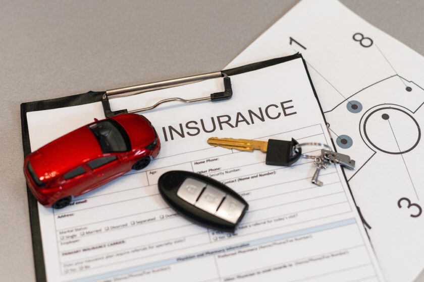 A red car model, a car key with a black fob, and a silver key are placed on top of a clipboard with an insurance form. The background includes a diagram on another paper.