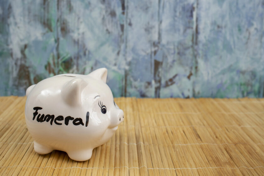 A white piggy bank with the word funeral written on it is placed on a bamboo mat. The background has a textured, abstract pattern in shades of blue and gray.