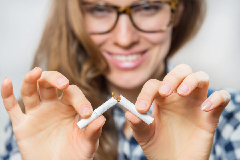 A woman wearing glasses smiles as she breaks a cigarette into two pieces. The focus is on her hands and the broken cigarette, symbolizing quitting smoking.