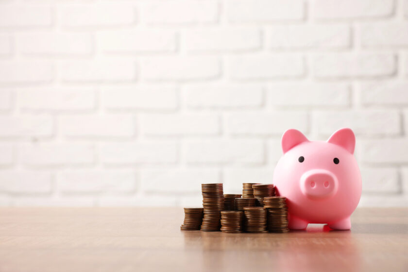 A pink piggy bank sits on a wooden surface next to stacked piles of coins. The background features a white brick wall.