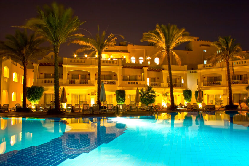 A luxurious resort at night with illuminated buildings reflecting in a tranquil swimming pool. The scene is adorned with palm trees and lounge chairs, creating an inviting and serene atmosphere.