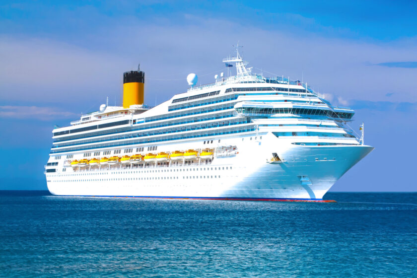 A large white cruise ship with multiple decks sailing on a calm blue sea under a clear sky. The ships imposing structure is complemented by its yellow lifeboats and the distinct black funnel stack on top.