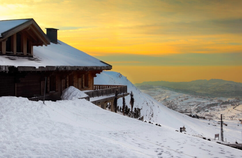 A wooden chalet with a snow-covered roof sits on a snowy slope, overlooking a vast landscape. The sky is painted with warm hues of orange and yellow from the setting sun, contrasting with the snow. The distant horizon is visible under the vibrant sky.