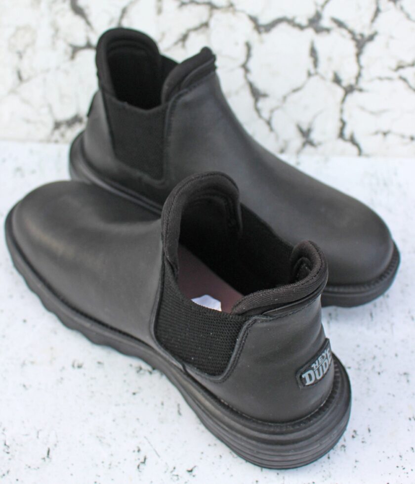 A pair of black slip-on ankle boots with elastic side panels and thick soles are placed on a textured white and black surface. A small tag on the heel displays the brand name.