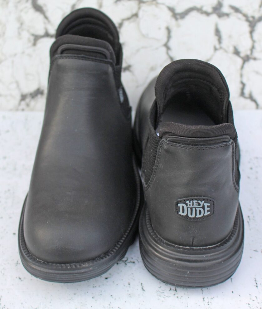 A pair of black leather slip-on shoes is shown. The right shoe displays a label on the heel with the text Hey Dude. The background is a white and gray textured surface.