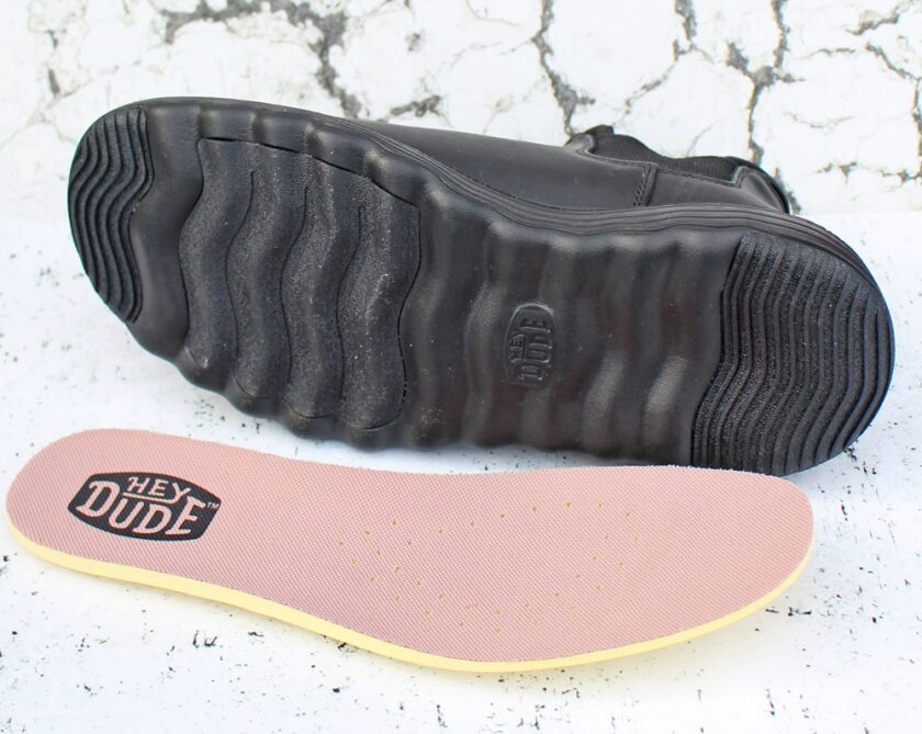 A black shoe lies on its side, showing a wavy-soled bottom and a branded insole reading Hey Dude in bold black letters. The shoe insole is pinkish with small perforations. The background is a cracked white surface.