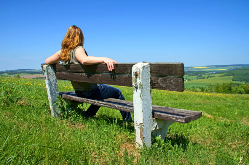 A person with long hair sits on a wooden bench atop a grassy hill, overlooking a vast landscape. The sky is clear and blue, with rolling hills and fields stretching into the distance. The scene is peaceful and sunny.